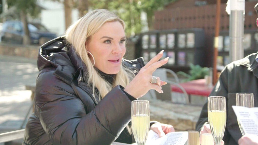 Heather Gay in a black puffer jacket gesturing over champagne at an outdoor table