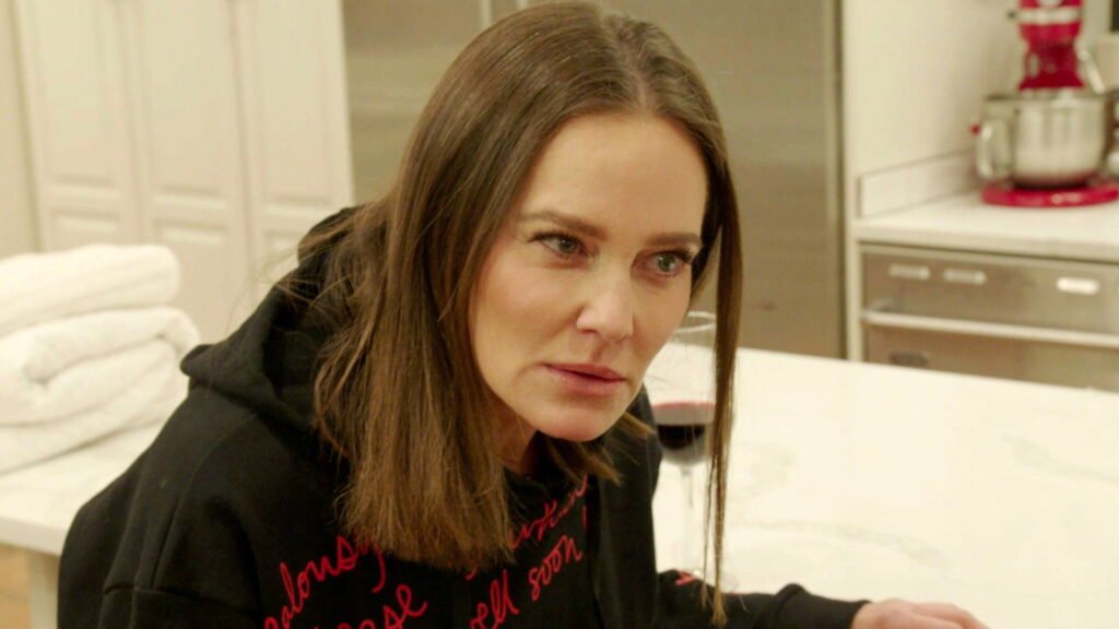Meredith Marks in a black hoodie at a kitchen counter, mid-conversation, a glass of red wine beside her