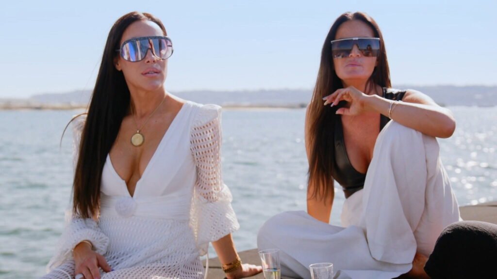 Angie Katsanevas in a white crochet dress and Lisa Barlow in oversized sunglasses, seated by the water in San Diego
