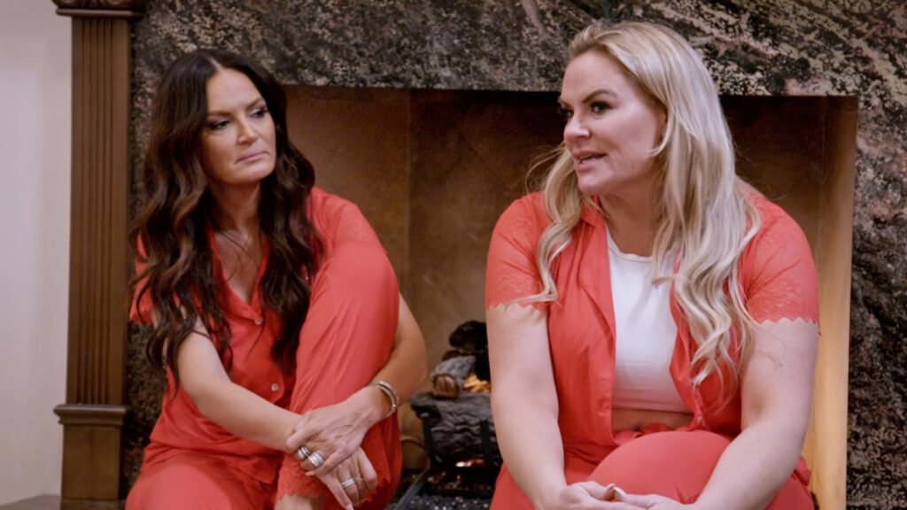 Lisa Barlow and Heather Gay in matching coral pajamas by a fireplace, mid-conversation