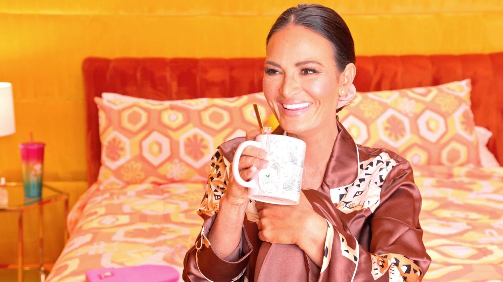 Lisa Barlow in satin pajamas on a bed at the Trixie Motel, smiling over a mug