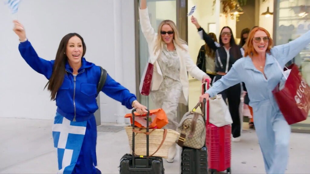 The cast arriving in Santorini with their luggage, one waving a small Greek flag