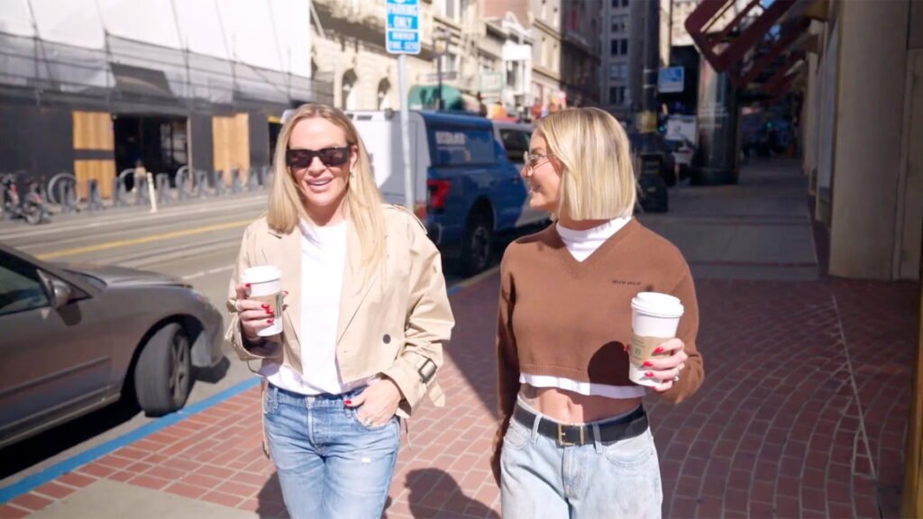 Two of the women walking down a city street with coffee cups